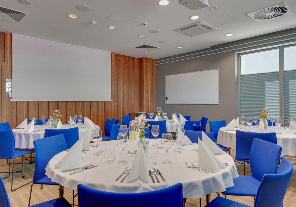 Banquet-style meeting room with round tables set for dining, white linens, folded napkins, blue chairs, floral centerpieces and projector screen.