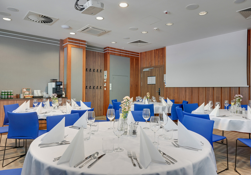 Conference dining room with round tables set with white linens, folded napkins, wine glasses, blue chairs, wood-paneled walls and projector screen.
