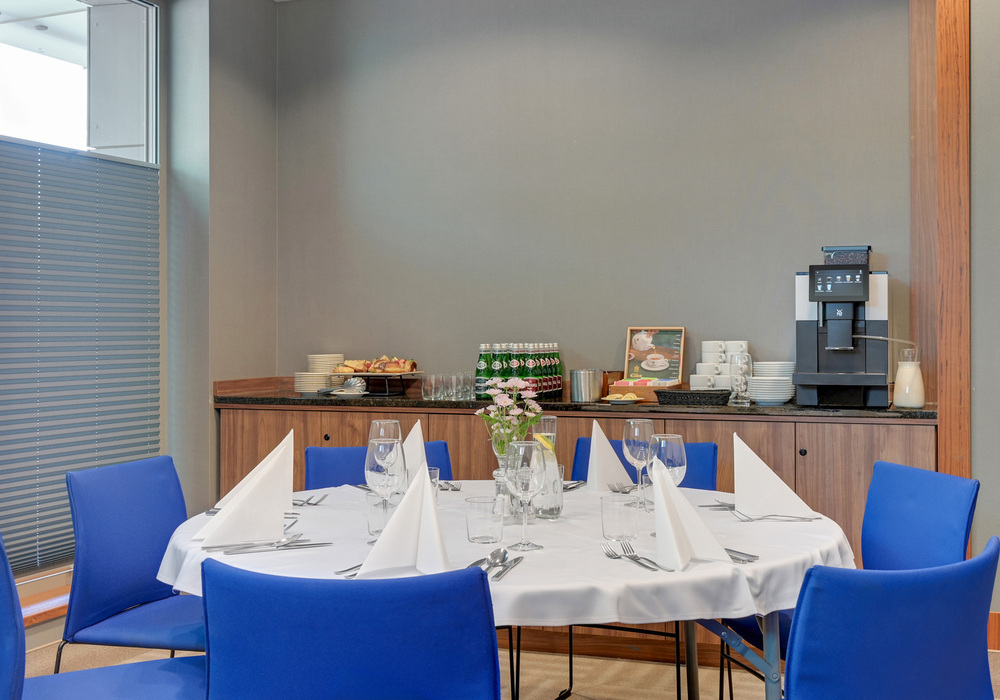 Round table set for meeting or private dining with white tablecloth, folded napkins, glassware, centerpiece flowers and coffee station with pastries.