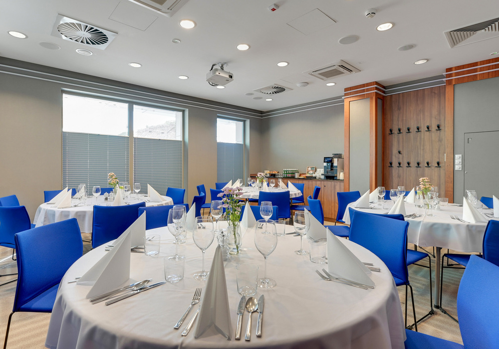 Bright banquet room with round tables in white linens, blue chairs, folded napkins, glassware, floral centerpieces and a coffee buffet
