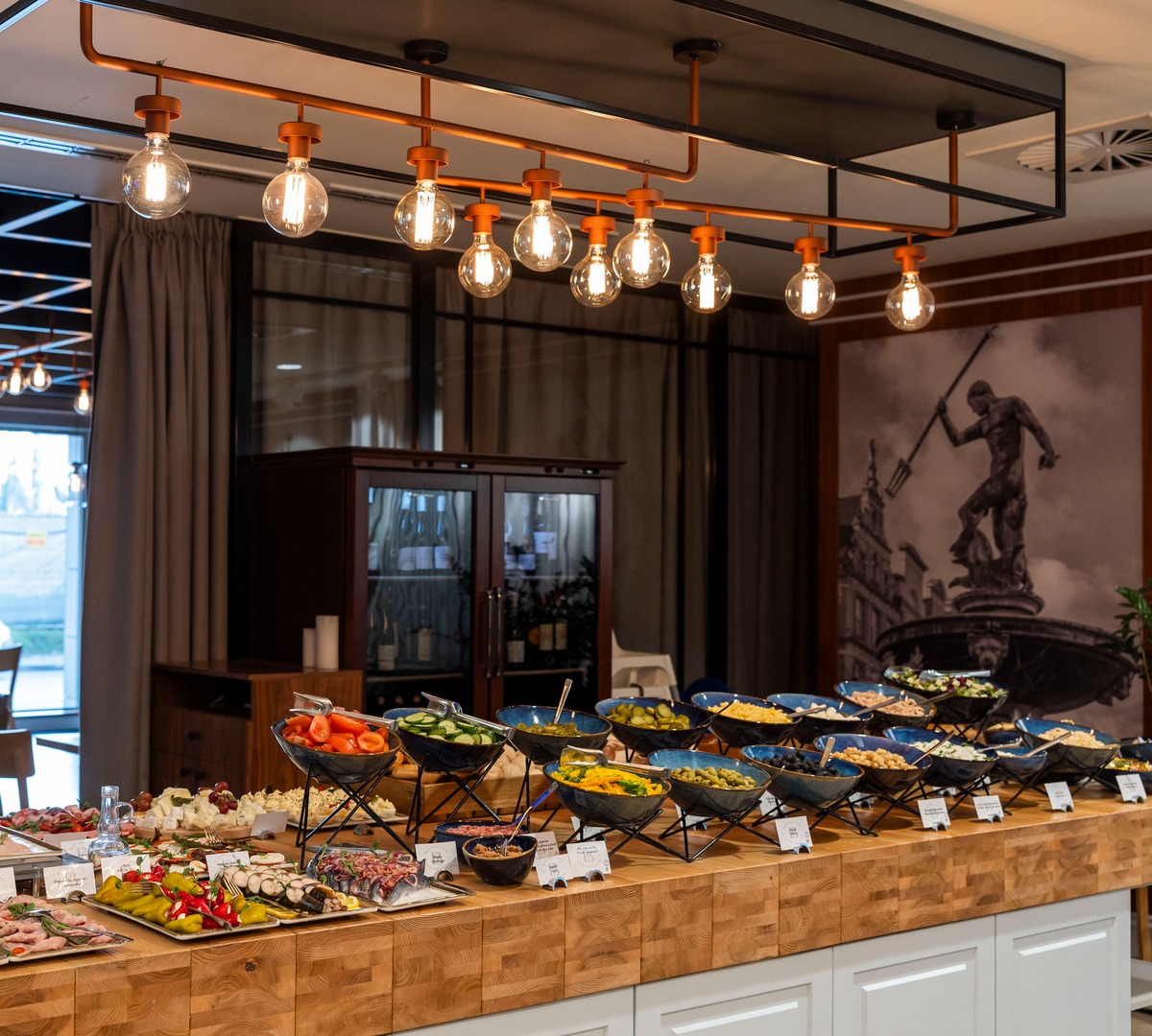 Hotellbuffé med pålegg, oster og friske salater i svarte skåler på tredisken under varme lyspærer.