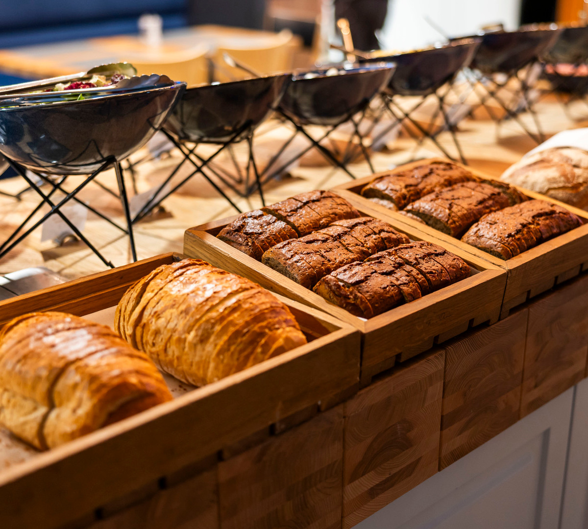 Gemütliches Frühstücksbuffet mit aufgeschnittenem Landbrot und Croissants in Holzschalen, Keramikschalen auf Metallgestellen