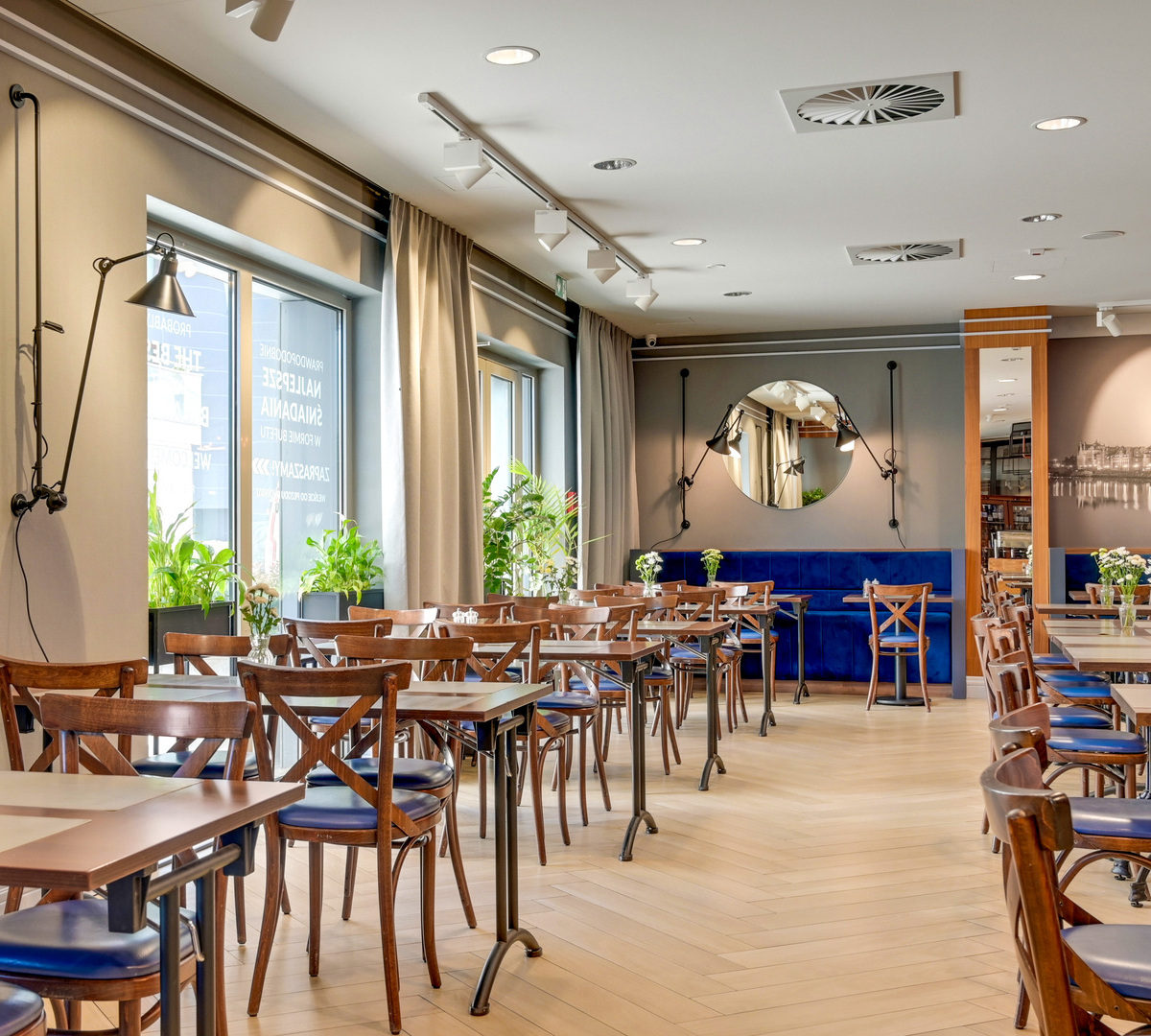 Jasna hotelowa jadalnia z drewnianymi stolikami, niebieską kanapą, regulowanymi lampami ściennymi, okrągłym lustrem i roślinami