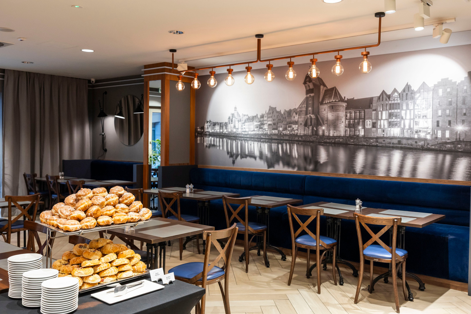 Breakfast buffet with stacked pastries and plates in a modern hotel dining room with navy banquette, wooden chairs and waterfront mural