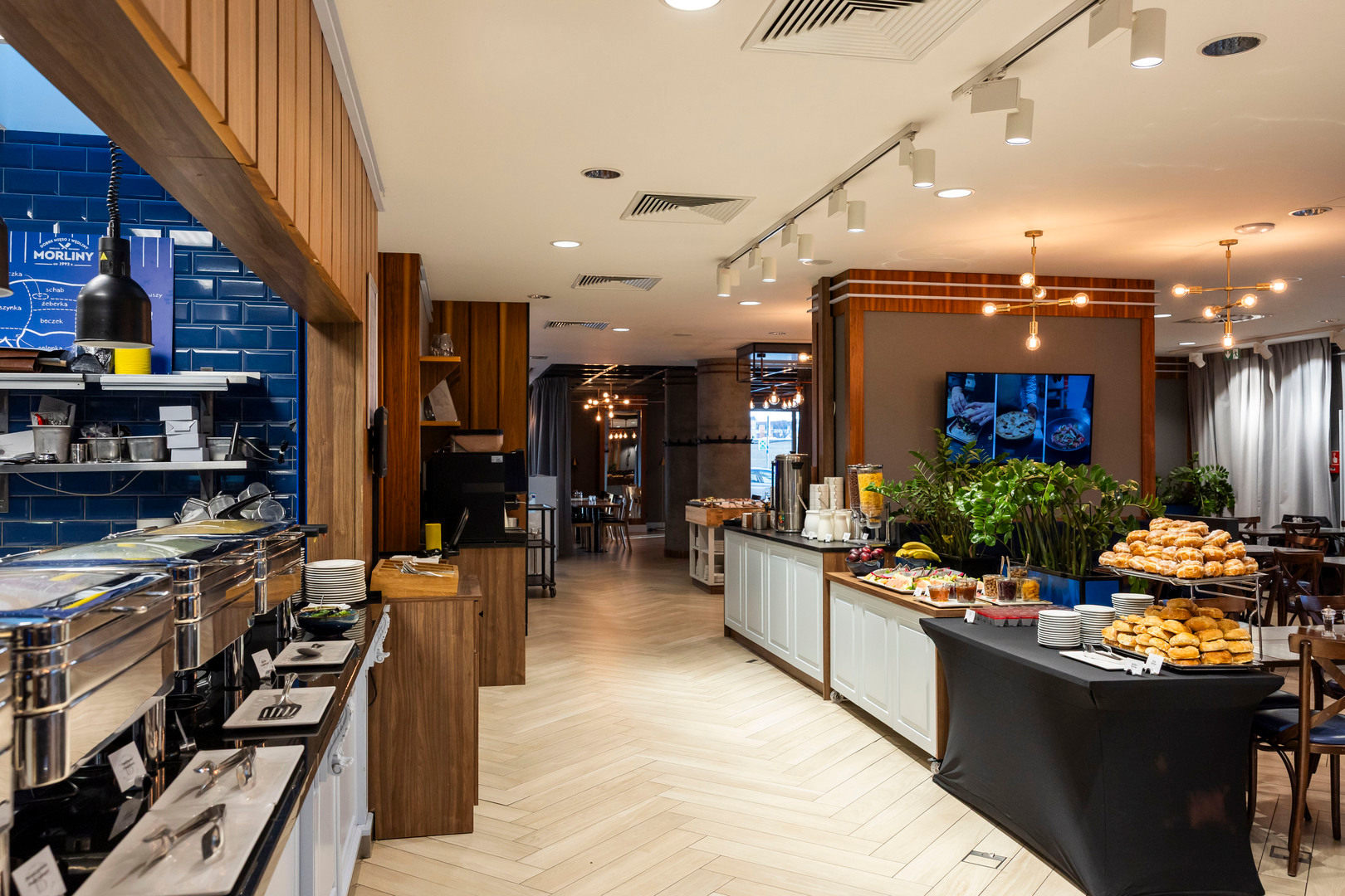 Breakfast buffet with chafing dishes, stacked pastries, cereal dispensers and coffee in a bright dining area with wood accents.