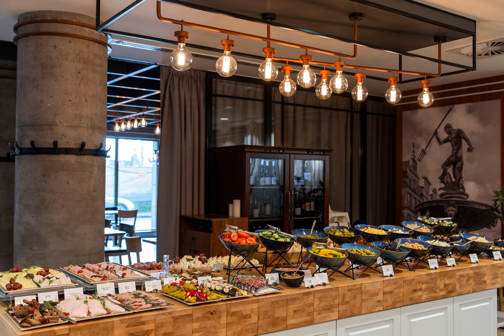 Generous hotel buffet of cold cuts, cheeses and salad bowls on a wooden counter beneath warm pendant lights