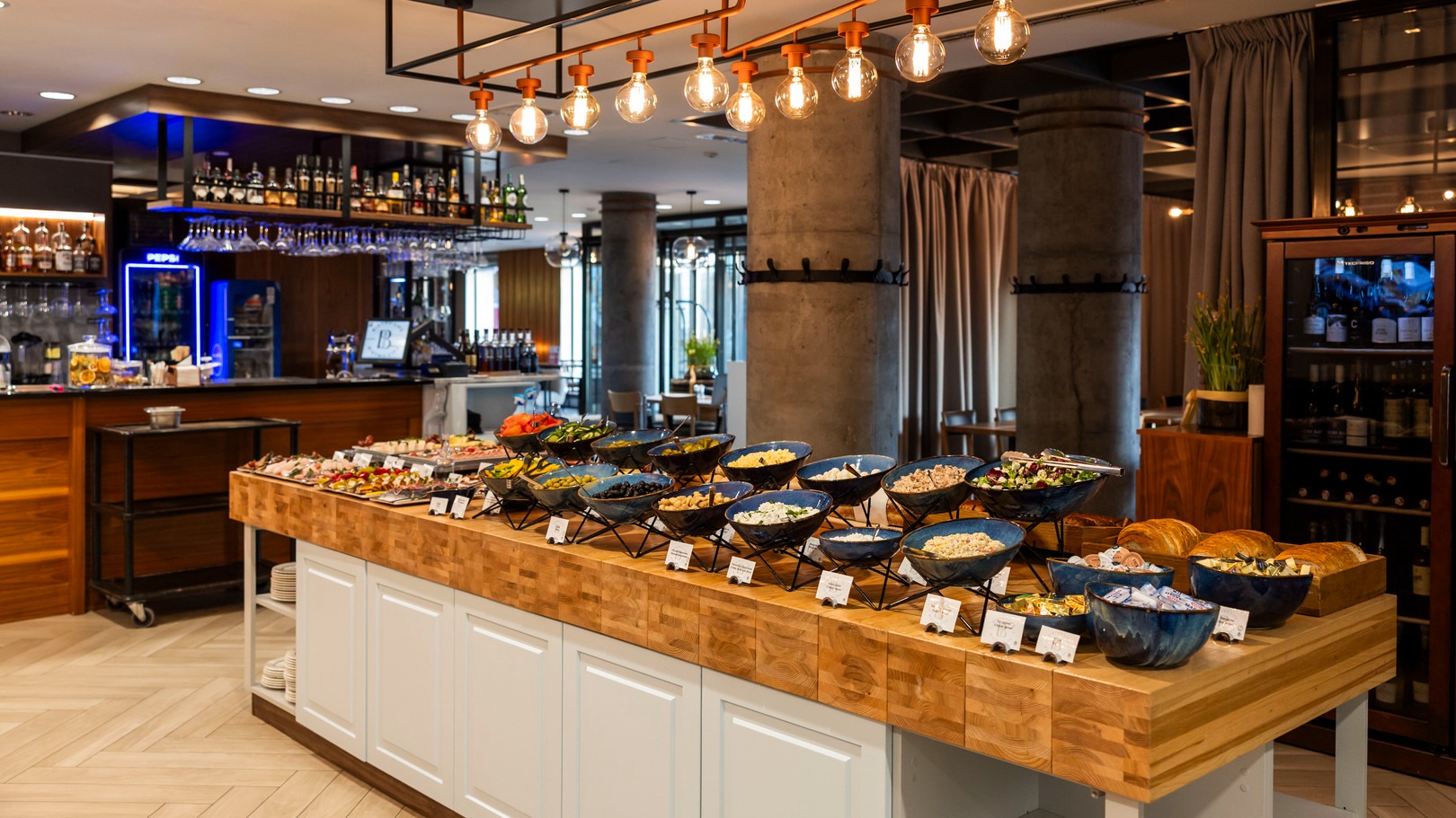 Hotelbuffet auf einer Holzinsel mit Schüsseln voller Salate, Brot und Beilagen, warmen Hängeleuchten und Bar im Hintergrund