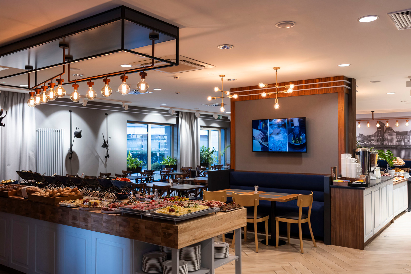 Breakfast buffet with breads, cheeses and cold cuts on a wooden island, coffee station, booth seating and warm pendant lights