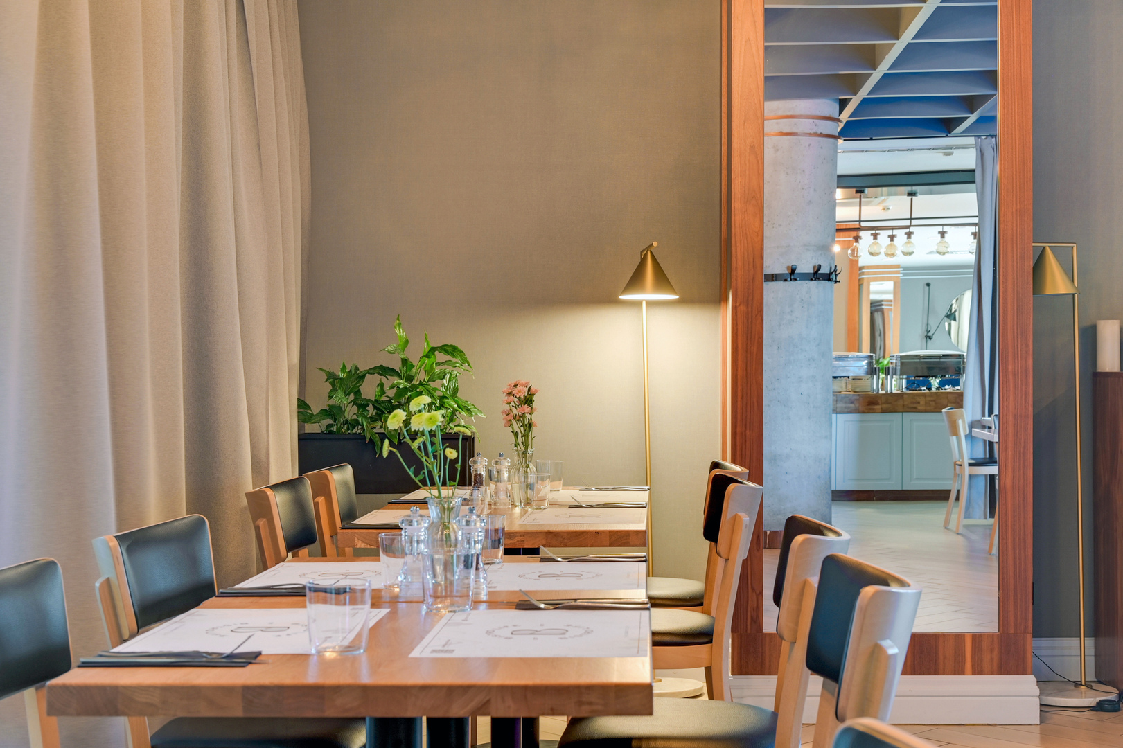 Contemporary hotel dining room with wooden tables set for guests, soft floor lamp, potted plants and mirrored view of buffet.