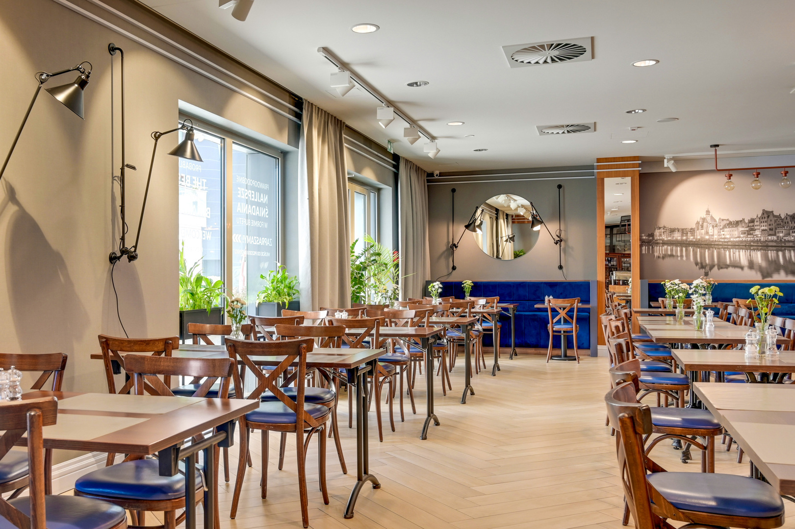 Bright hotel dining room with wooden tables, blue banquette seating, adjustable wall lamps, round mirror, plants and flower vases