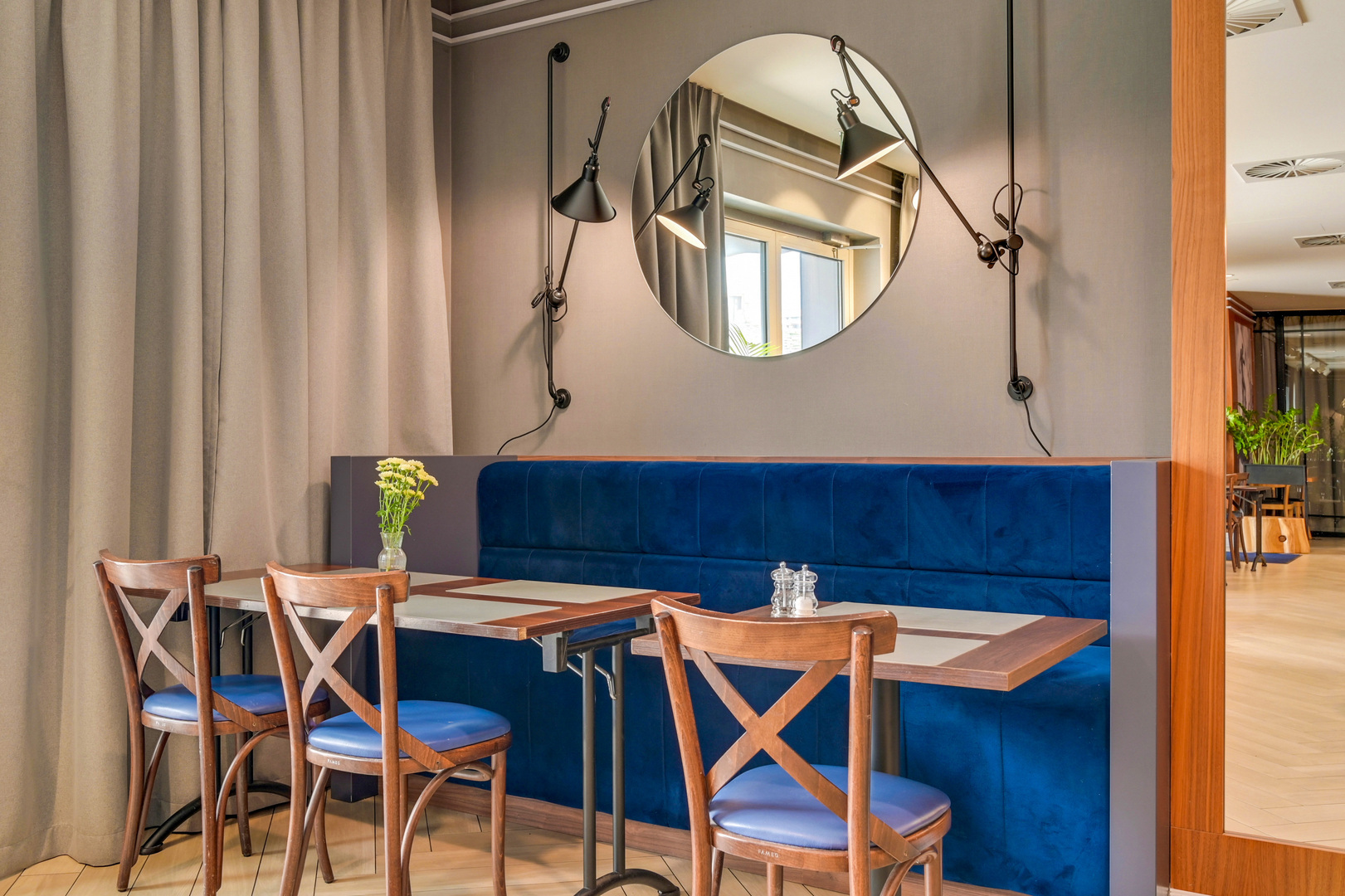 Cozy hotel dining nook with blue velvet banquette, wooden chairs, round mirror, wall lamps and flower vase