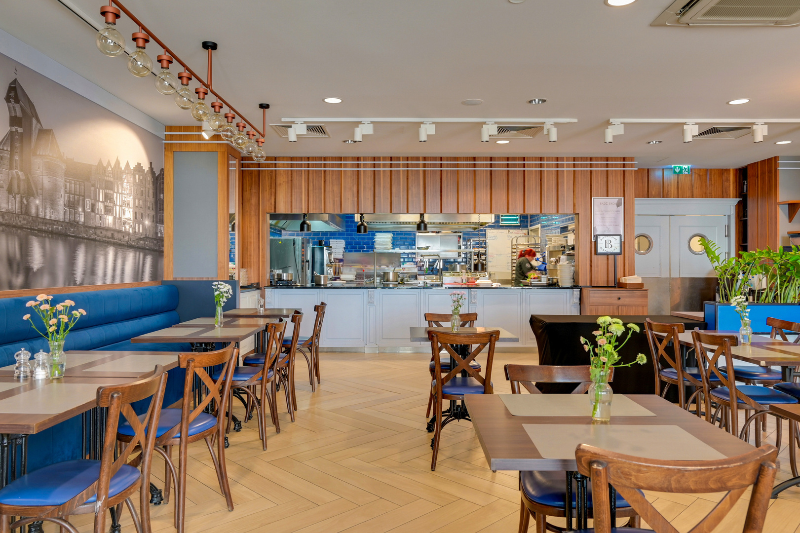 Hotel dining room with open kitchen, wood-paneled wall, blue banquette seating, wooden tables and fresh flowers