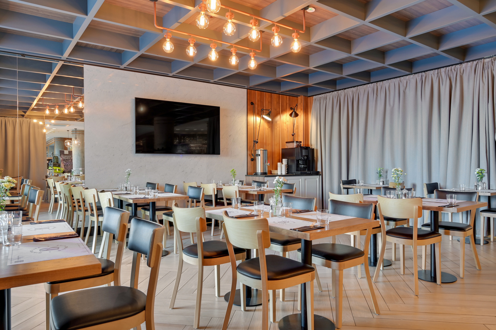 Modern hotel restaurant with wooden tables and chairs set for dining, filament pendant lights, wall TV and coffee station