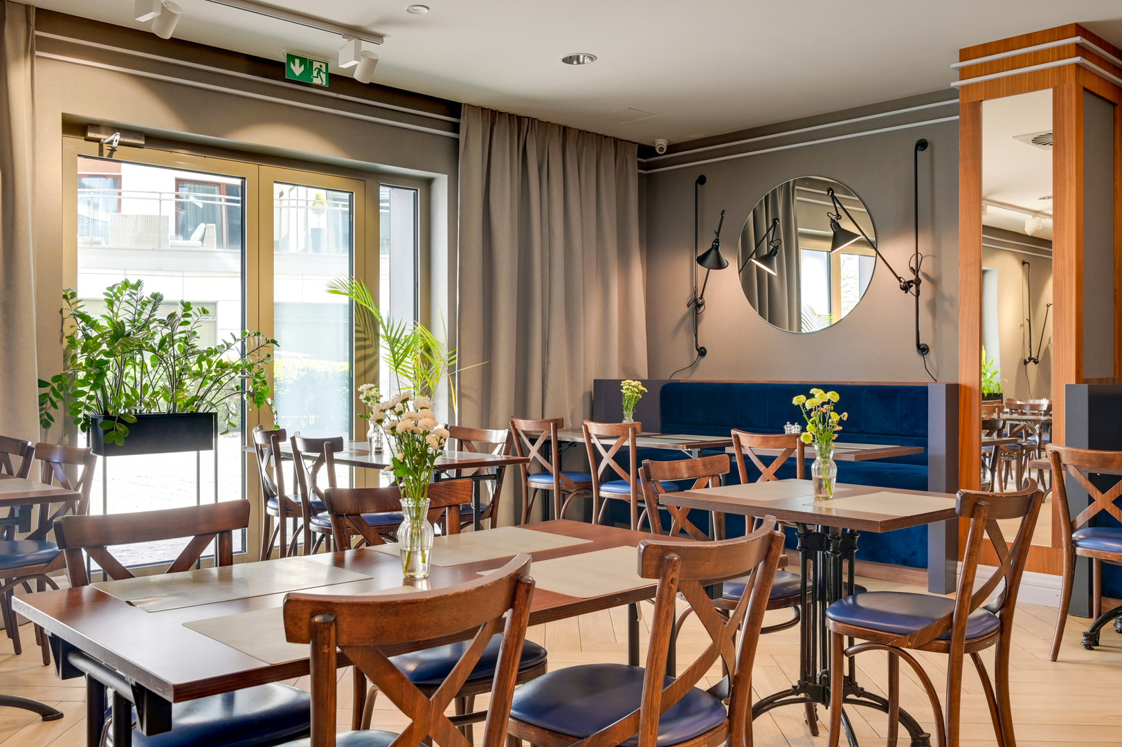 Hotel dining area with wooden tables and chairs, blue banquette seating, vases of fresh flowers, large windows and indoor plants