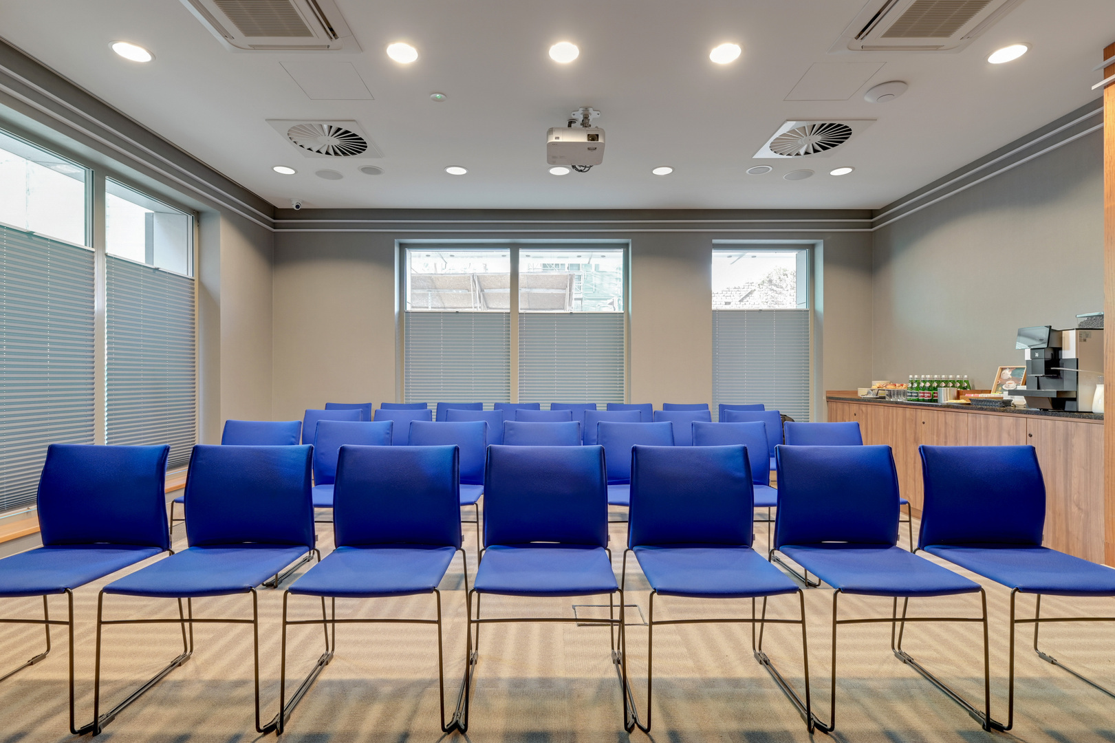 Konferensrum med rader av blå stolar, takprojektor, kaffe- och snacksstation, persienner och modern, neutral inredning