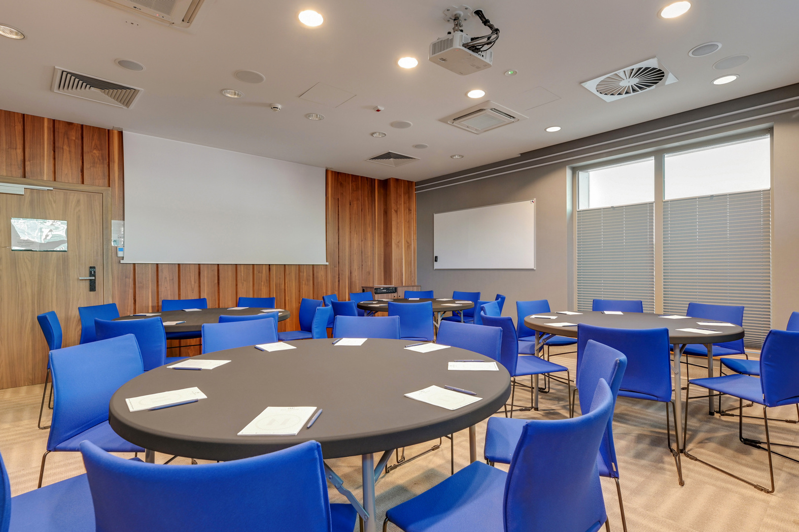Konferensrum med runda bord, blå stolar, takmonterad projektor, whiteboard och träpanelvägg