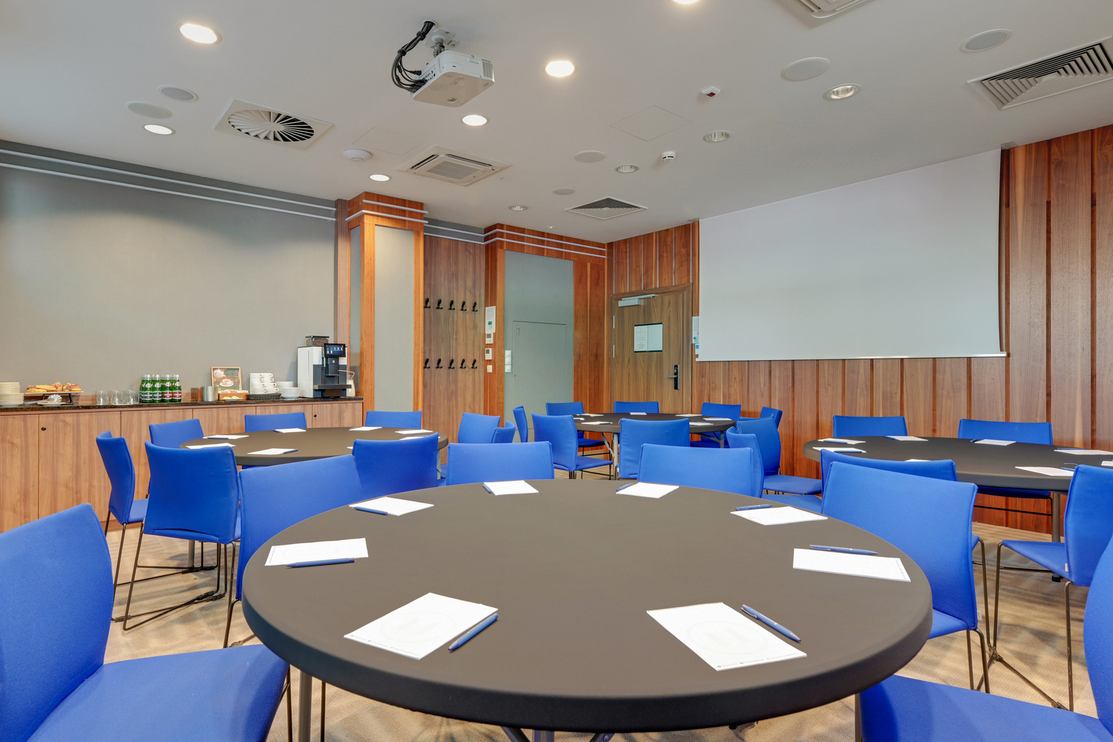 Konferensrum med runda bord, blå stolar, anteckningsblock och pennor, projektor och duk, kaffestation och träpanel på väggarna