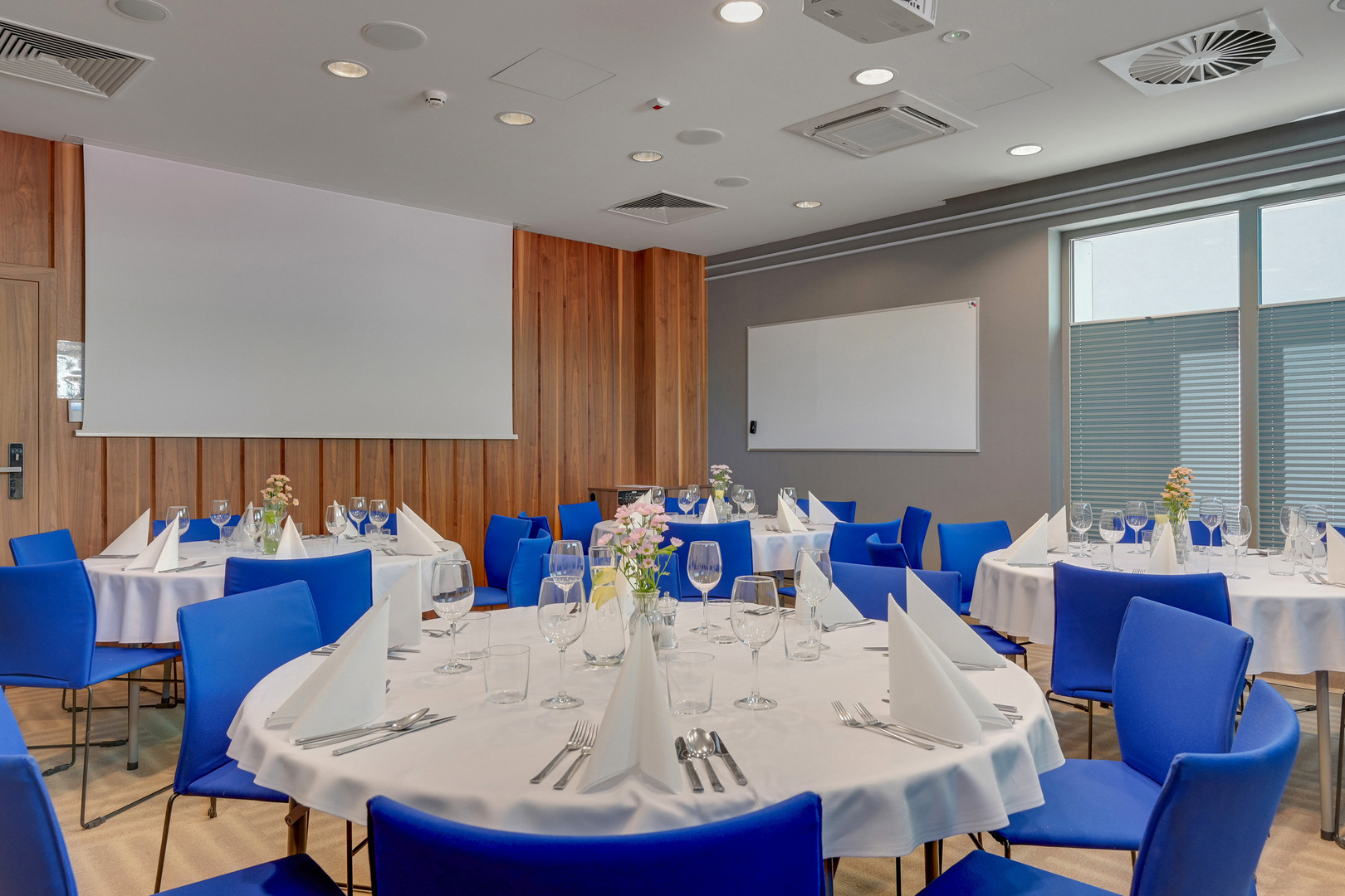 Banquet-style meeting room with round tables set for dining, white linens, folded napkins, blue chairs, floral centerpieces and projector screen.