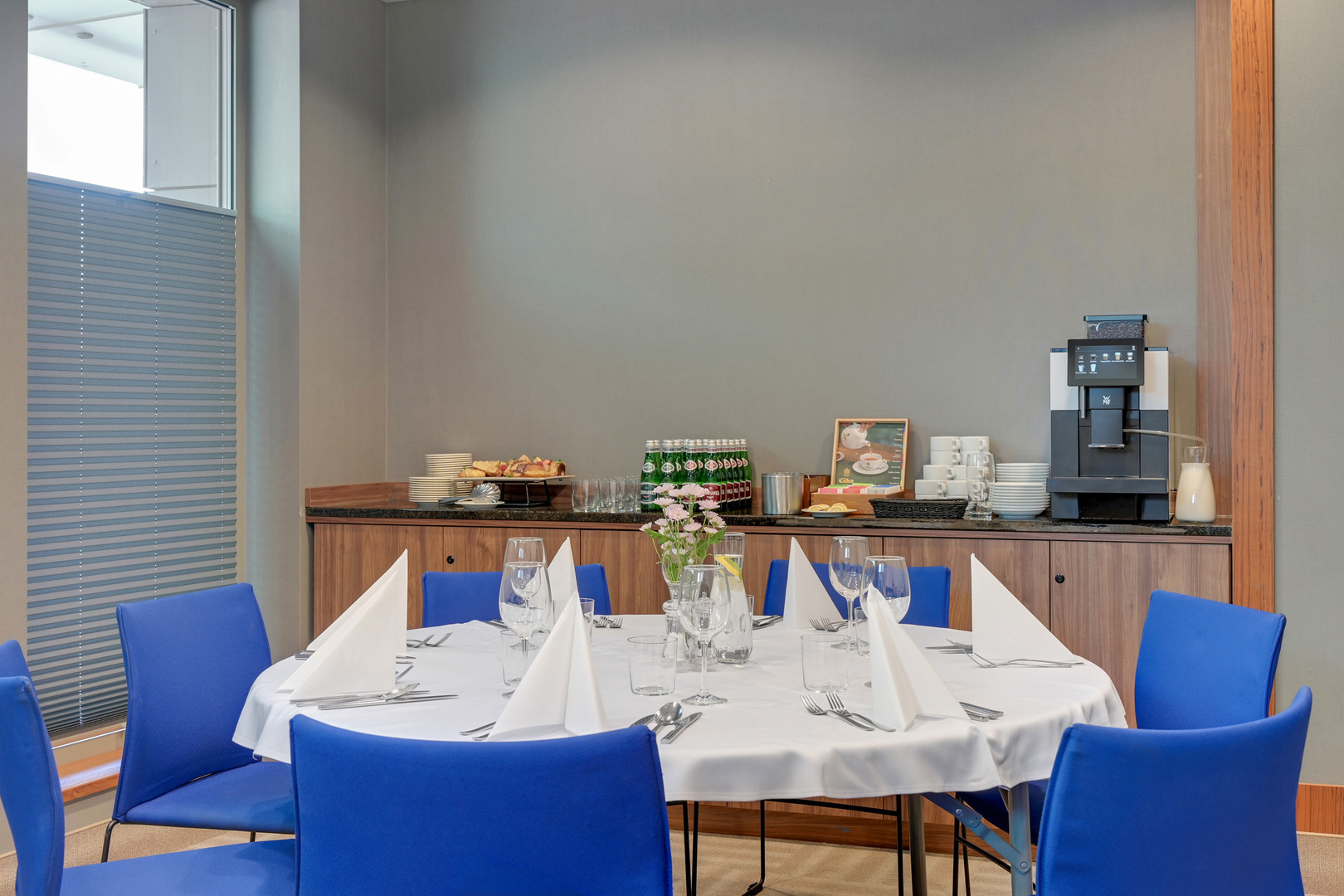 Runt bord dukat för möte eller privat middag med vit duk, vikta servetter, glas, blommor och kaffestation med maskin och bakverk