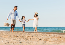 Familie geht barfuß am Sandstrand eines Strandhotels, Eltern halten Kind an den Händen, Picknickkorb und Meerblick