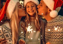 Eltern küssen ihr lächelndes Kind in Weihnachtsmützen, halten Wunderkerzen in warmem, gemütlichem Festtagsambiente