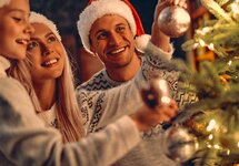 Familie mit Weihnachtsmützen hängt glitzernde Kugeln an einen warm beleuchteten Tannenbaum, gemütliche Festtagsstimmung