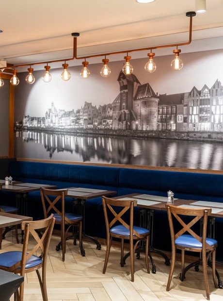 Frokostbuffé med stabler av bakverk og tallerkener i moderne hotellspisesal med mørk blå sofa, trestoler og elvebreddemotiv