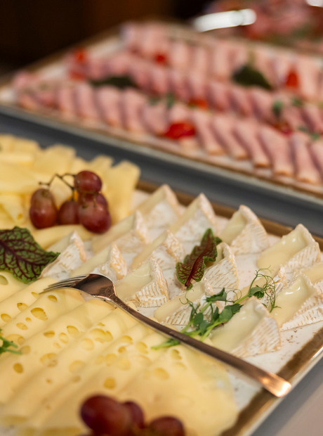 Buffetplatte mit Scheiben Schweizer Käse und Brie-Stücken, garniert mit Trauben und Kräutern; im Hintergrund aufgerollter Schinken