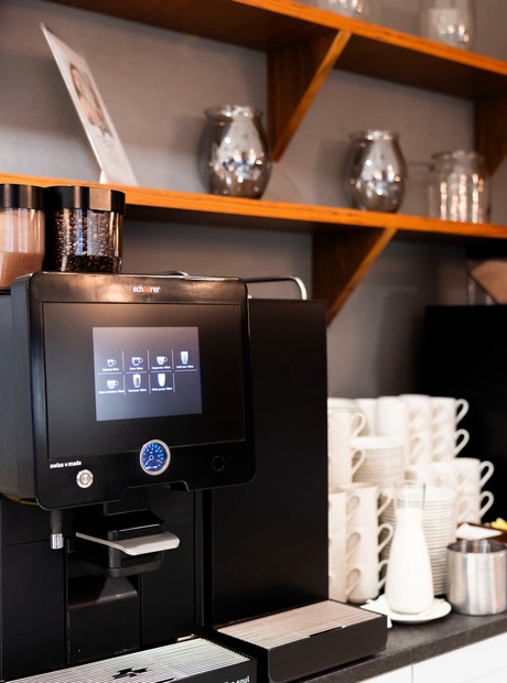 Selbstbedienungs-Kaffeeplatz mit zwei schwarzen Espressomaschinen, gestapelten weißen Tassen, Milchkanne, Zucker und Holzregalen mit Gläsern