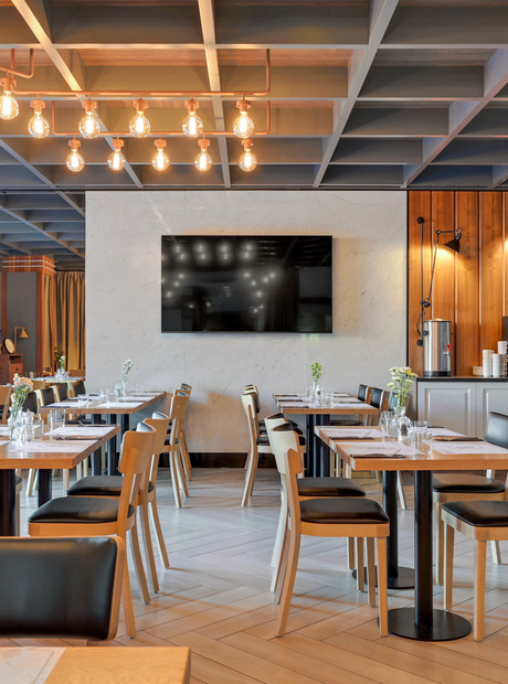 Contemporary hotel dining room with wooden tables set for meals, black leather chairs, hanging pendant lights, TV and coffee station.