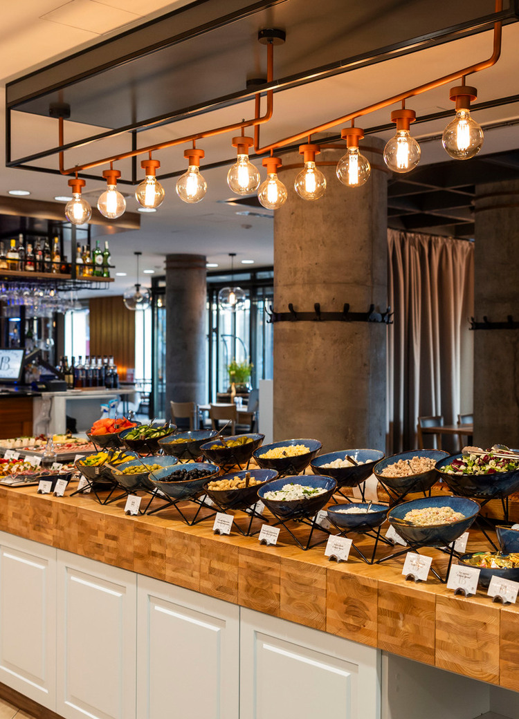 Hotel buffet spread on wooden island with bowls of salads, breads and condiments, warm pendant lights and a bar in background