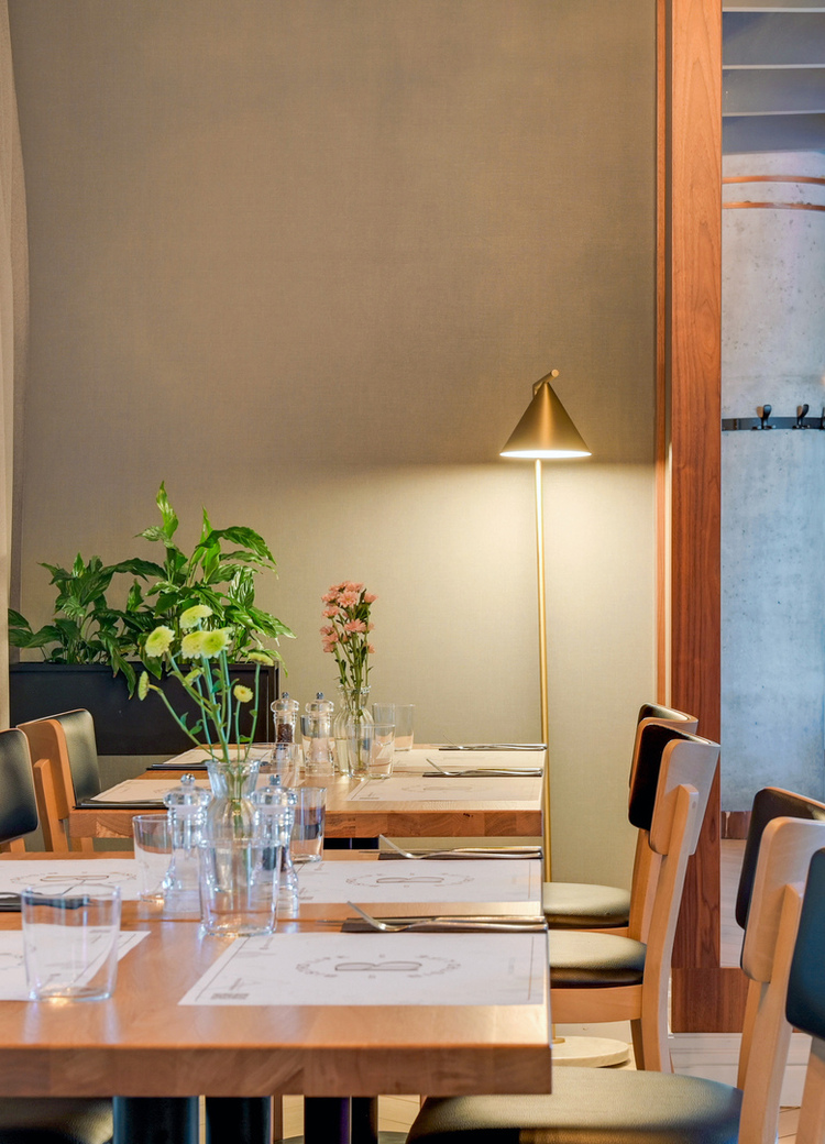 Contemporary hotel dining room with wooden tables set for guests, soft floor lamp, potted plants and mirrored view of buffet.