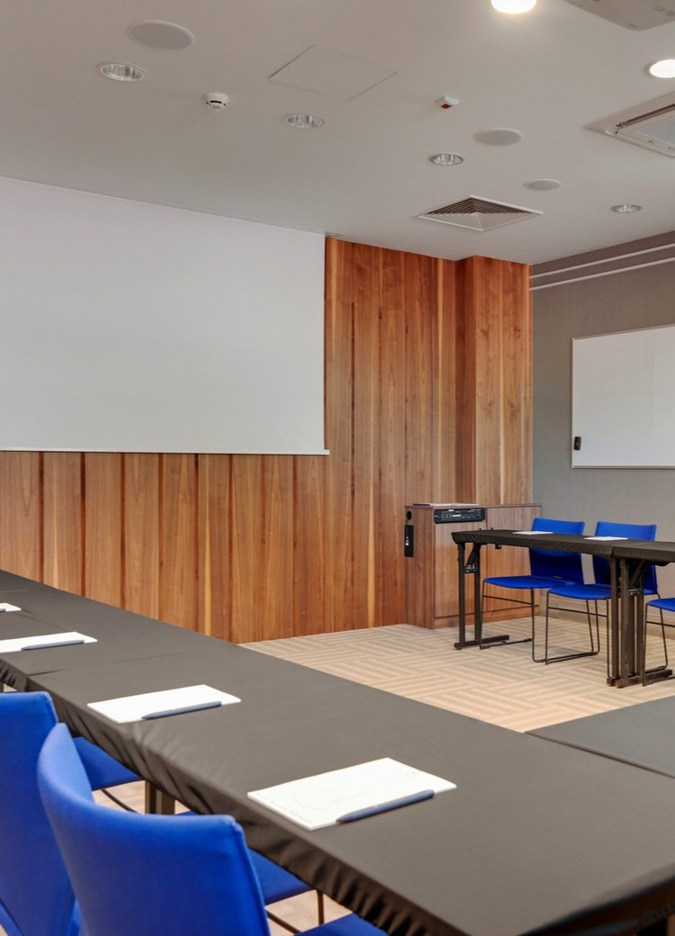 U-shaped meeting room with blue chairs, black-covered tables, projector screen, whiteboard and wood-paneled wall