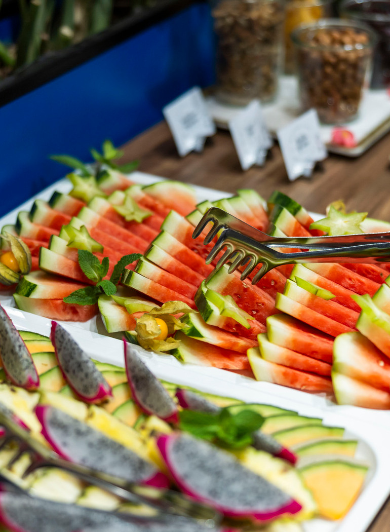 Buntes Obstbuffet mit aufgeschnittener Wassermelone, Drachenfrucht und Melone, Minze und Servierzange
