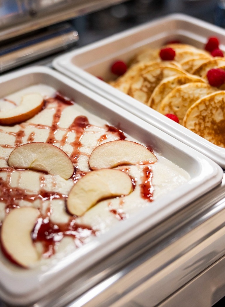 Frühstücksbuffet mit goldenen Pfannkuchen und Himbeeren, daneben cremiger Grießbrei mit Apfelscheiben und Beerensauce