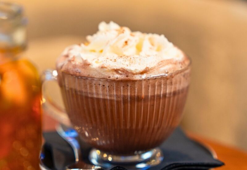 Clear glass mug of hot chocolate topped with whipped cream on a wooden table, served with a spoon on a black napkin.