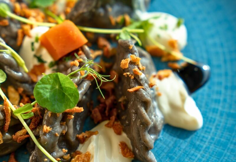 Black dumplings on a blue plate served with creamy sauce, crispy toppings, and fresh green herbs.