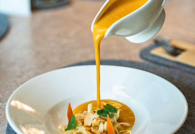 Smooth pumpkin soup being poured into a white bowl garnished with vegetables, almonds, and fresh herbs on an elegant restaurant table.