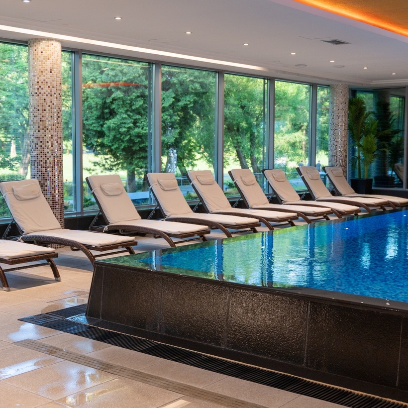 Przyhotelowy basen kryty z szerokimi leżakami i panoramicznymi oknami na zielony ogród.
