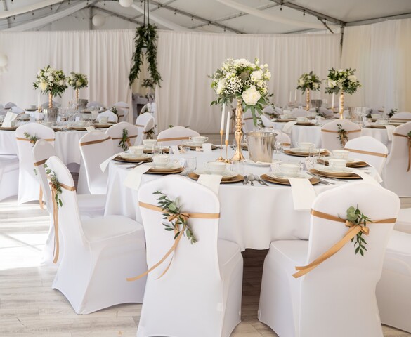 Elegant wedding reception in a white tent with round tables, gold accents, floral centerpieces and draped chair sashes.