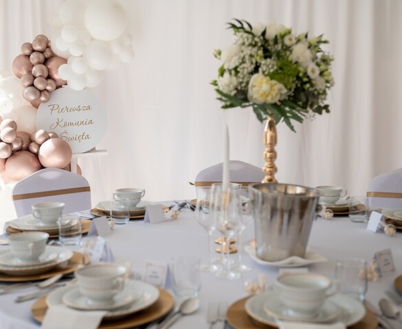 Elegant round table set for a First Communion with white china, gold chargers, tall floral centerpiece and balloon garland.