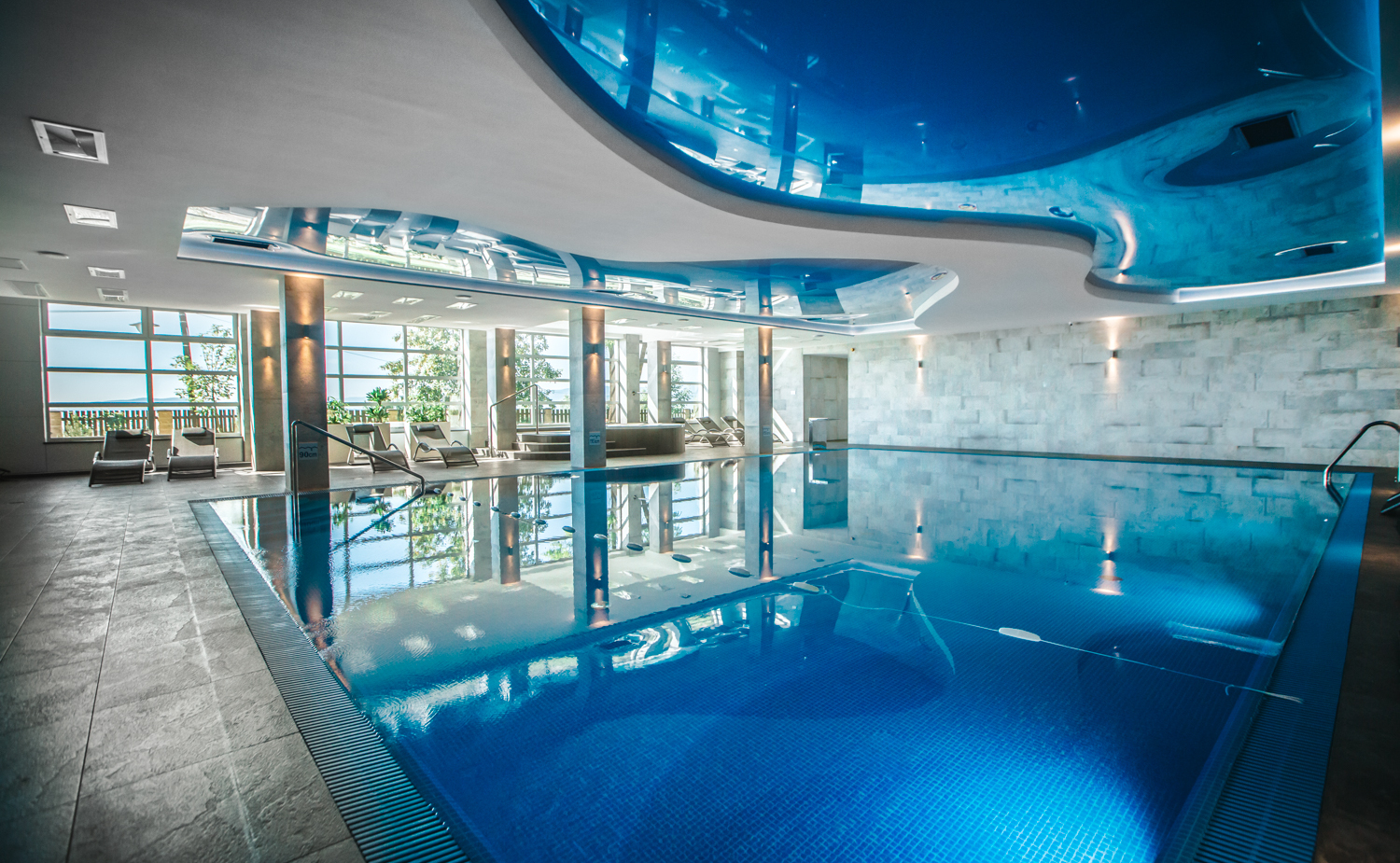 Kryty basen hotelowy z spokojną błękitną wodą, giętym lustrzanym sufitem, dużymi oknami i leżakami na słonecznym tarasie