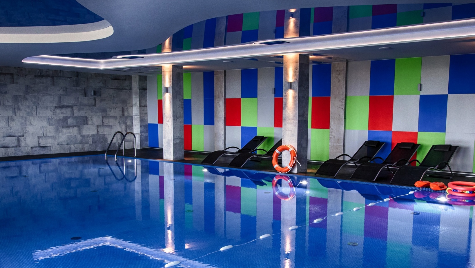 Kryty basen hotelowy z niebieską mozaiką, leżakami, kolorowymi panelami ściennymi, nastrojowym oświetleniem i kołem ratunkowym