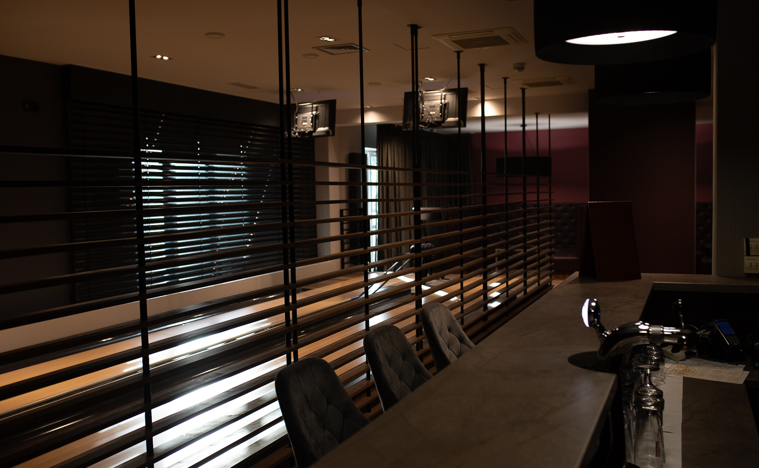 Moderne Hotelbar mit gepolsterten Hockern, Zapfhahn und Gläsern, Holzlamellen-Trennwand zeigt gedämpften Lounge-Bereich mit Fernsehern.