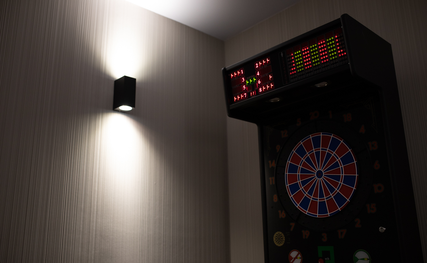 Elektronische Dartscheibe mit beleuchtetem LED-Display neben einem Wandleuchter auf strukturierter Tapete in einer Spielzone