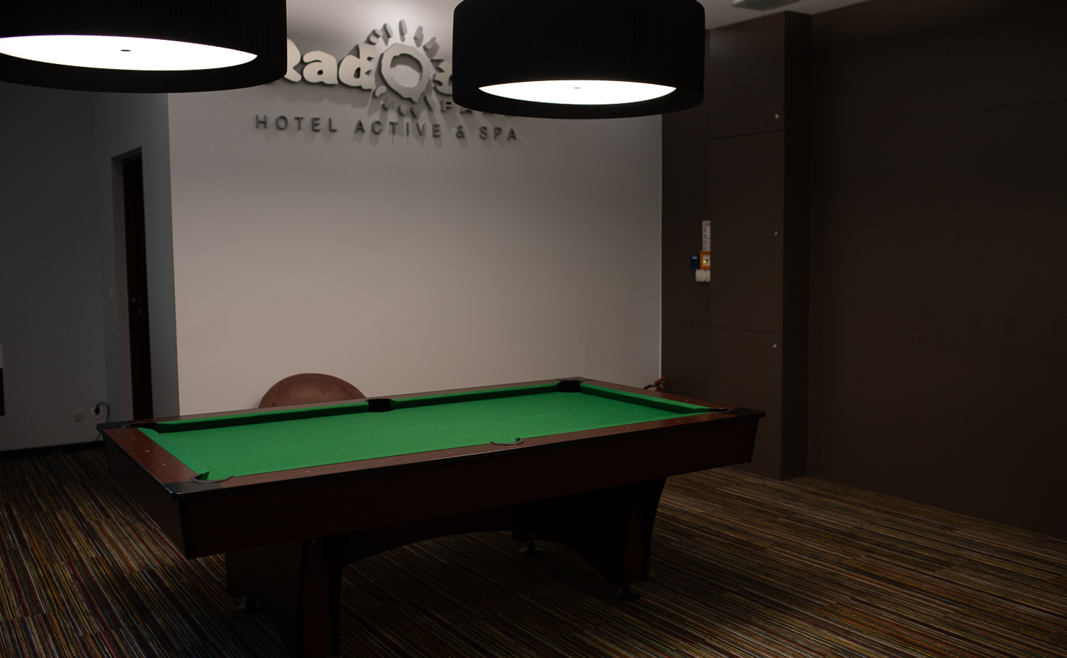 Hoteleigener Spielraum mit grünem Billardtisch, modernen Hängeleuchten, gestreiftem Teppich und Sessel unter dem Schriftzug 'Hotel Active & Spa'