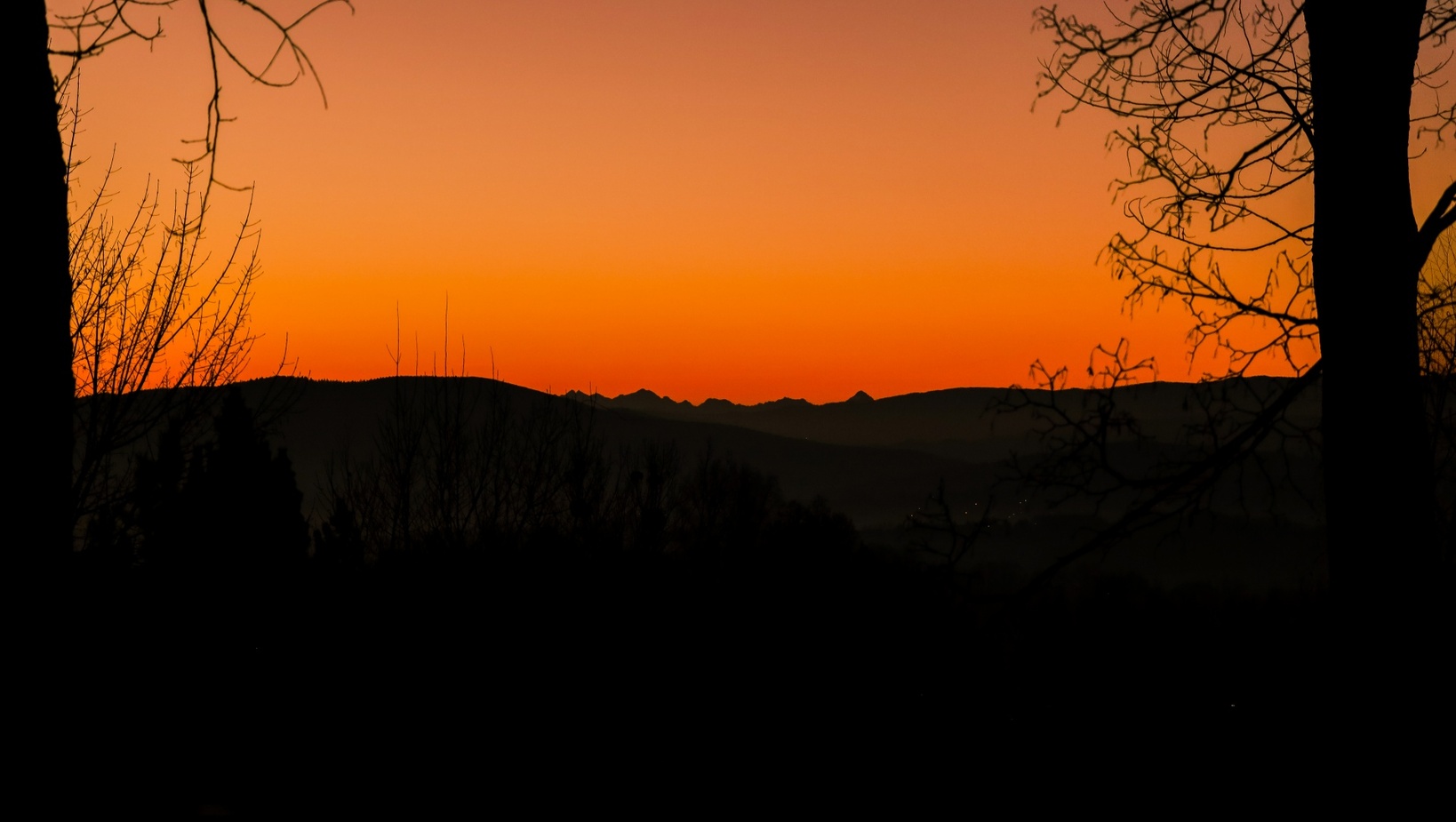 Západ slnka nad viacerými horskými hrebeňmi, oranžová obloha, stromy v siluete rámujú údolie.