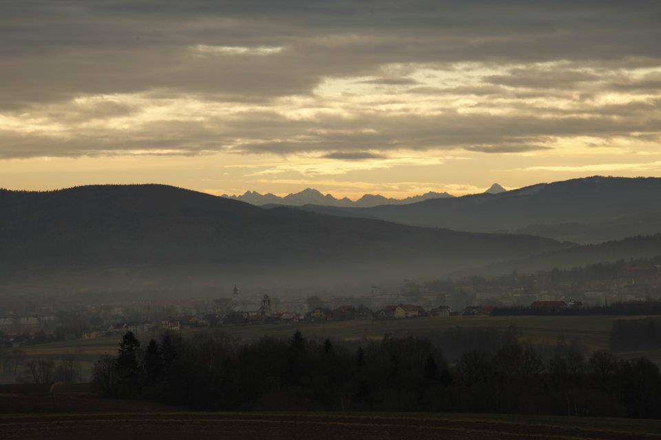 Hmlistá panoráma údolia z terasy: dedina s kostolnými vežami, vlnité kopce a vzdialené horské štíty v zlatom úsvite