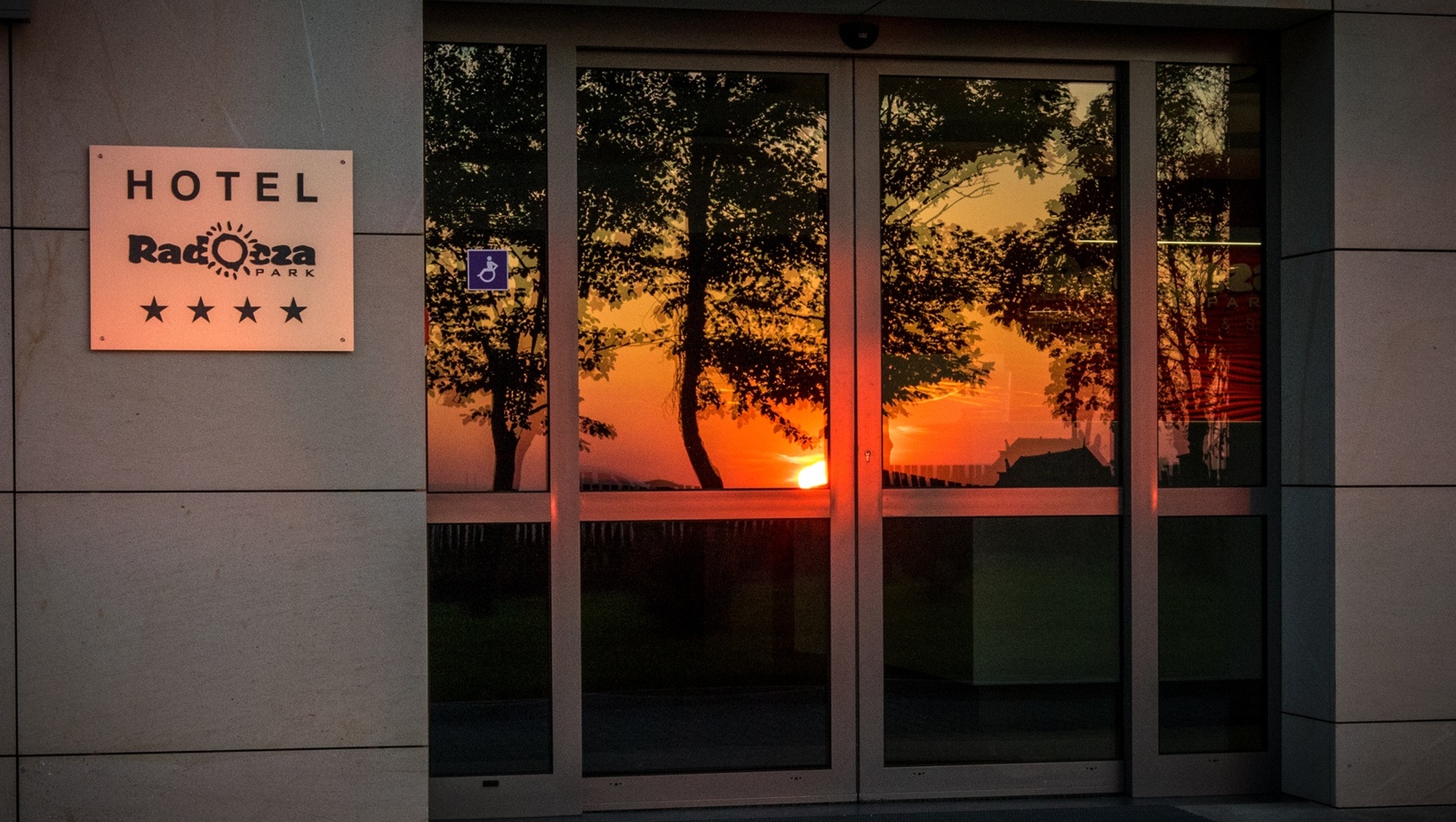 Vstup do hotela s presklenými dverami odrážajúcimi teplý západ slnka a stromy; tabuľa so štyrmi hviezdičkami a symbolom prístupnosti.