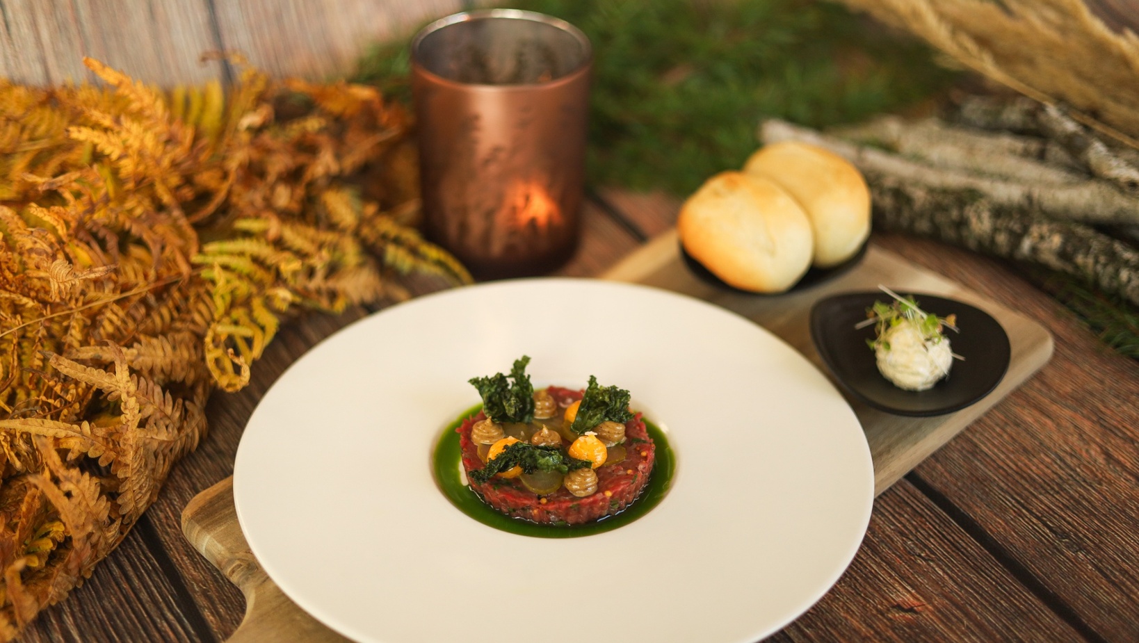 Hovězí tatarák s křupavým kapustou a bylinkovým olejem, podávaný s houskami a máslem na rustikální desce u svíčky